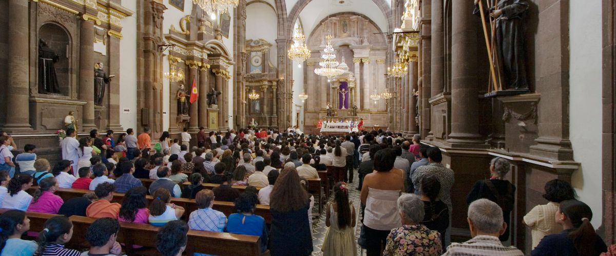 Crowded church congregation attending Mass inside an ornate cathedral with chandeliers and altar at the front.