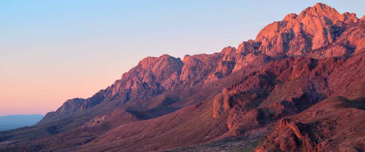 Mountain range glowing with pink sunset light over rocky desert landscape.