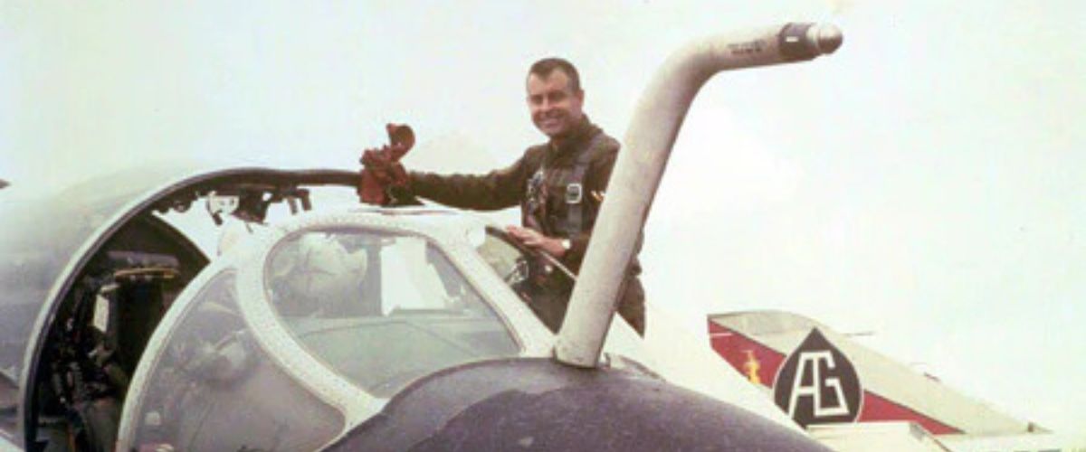 **Alt text:** Smiling pilot standing in an open fighter jet cockpit, holding a small teddy bear.