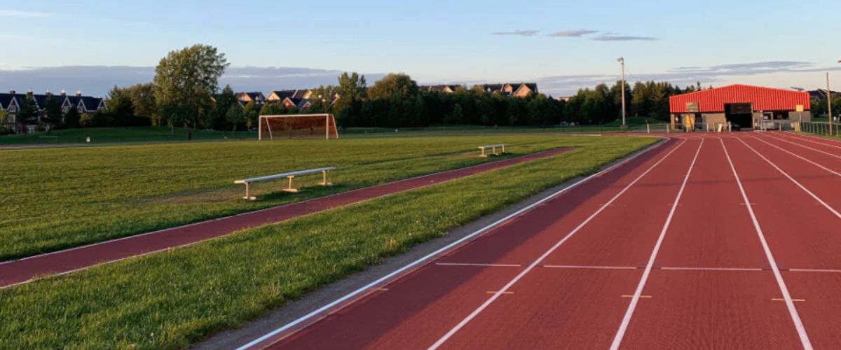 Red running track beside a grassy soccer field with goalposts, benches, and a small red building under a clear evening sky.