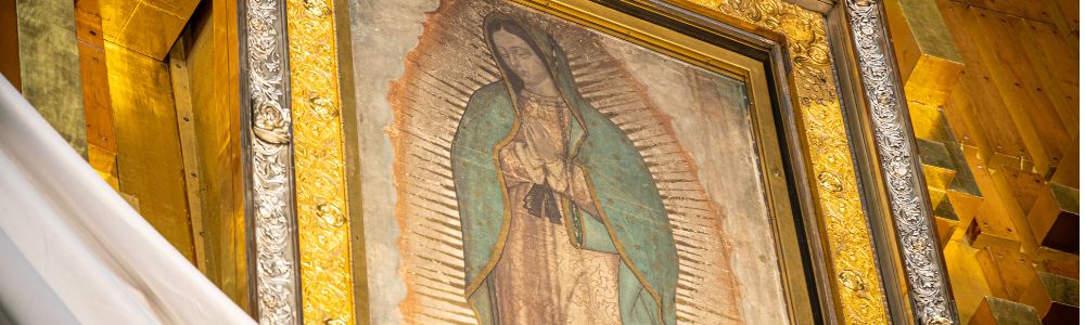 Framed image of Our Lady of Guadalupe with a gold ornate border displayed inside a church.