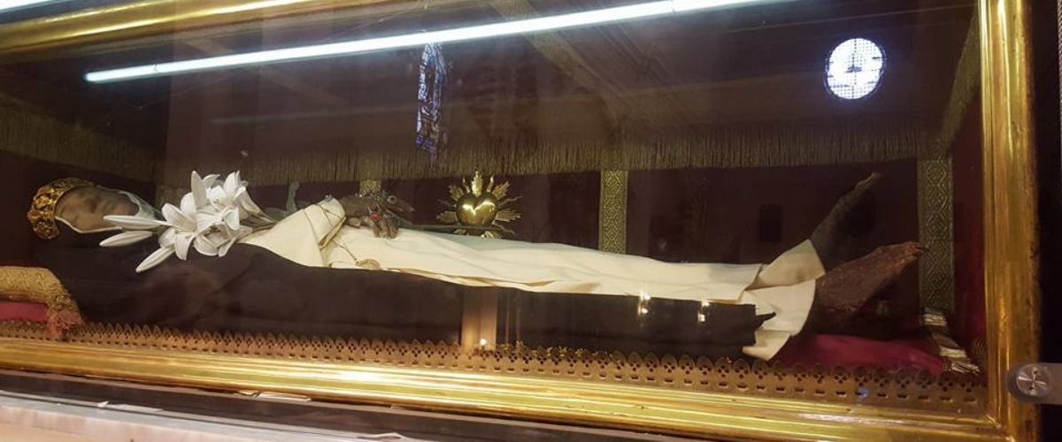 A preserved body displayed in a glass case, adorned with a crown and white lilies. The background features decorative elements, including a golden heart, under soft lighting.