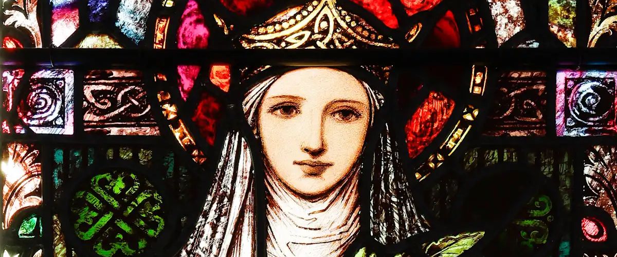A stained glass window depicting St. Brigid, shown wearing a crown and holding a lamp, surrounded by vibrant floral patterns. She is dressed in richly detailed robes, symbolizing her spiritual significance and legacy.