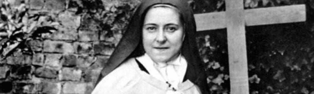 Black-and-white photo of St. Therese in a habit holding flowers, standing beside a wooden cross outdoors.