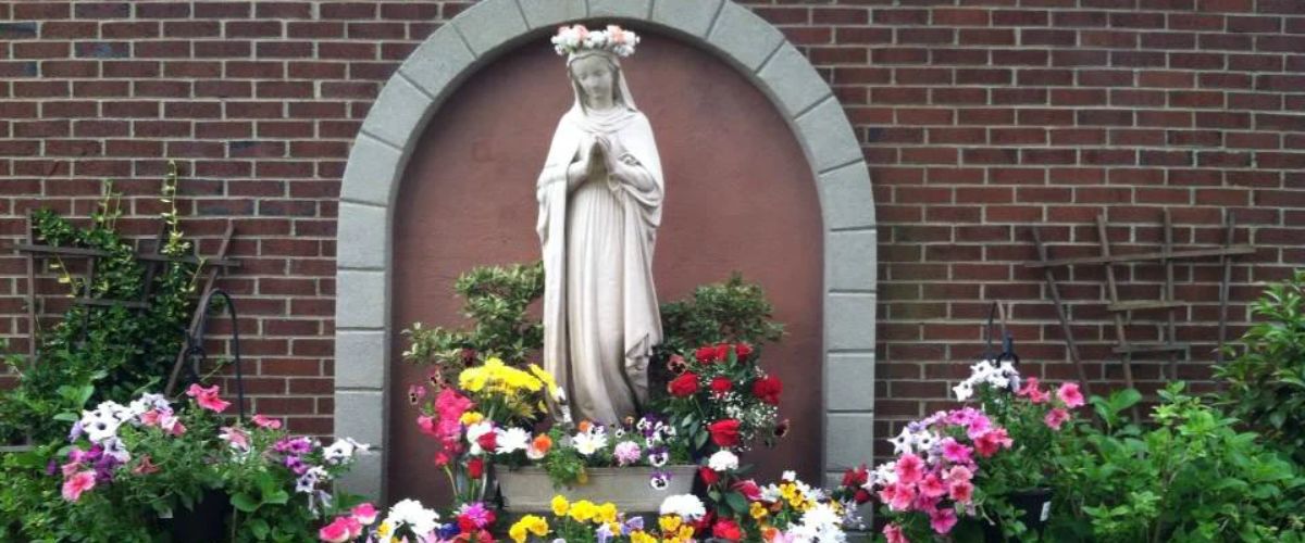 Statue of the Virgin Mary in a garden shrine surrounded by colorful flowers against a brick wall