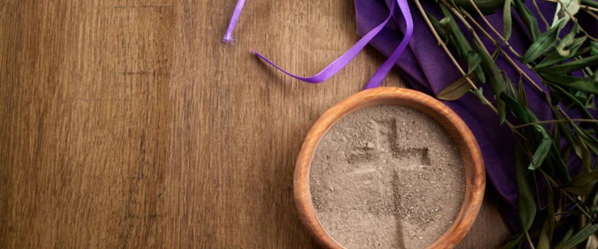 Bowl of ashes with a cross, draped in purple cloth and palm branches on a wooden surface.