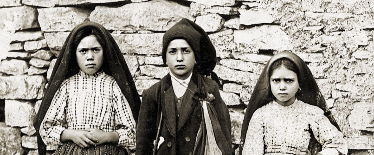 Black-and-white photo of the three Fatima child visionaries standing in front of a stone wall