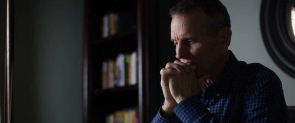 Man sitting at a desk, hands clasped in thought, with an open book and a cup nearby.