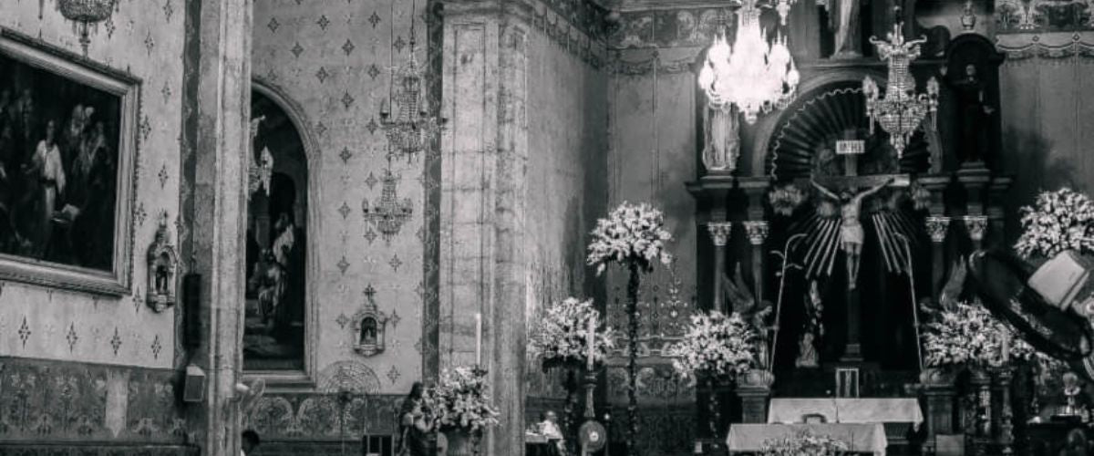 Black-and-white photo of a church interior with worshippers seated, ornate arches, chandeliers, and an altar at the front.