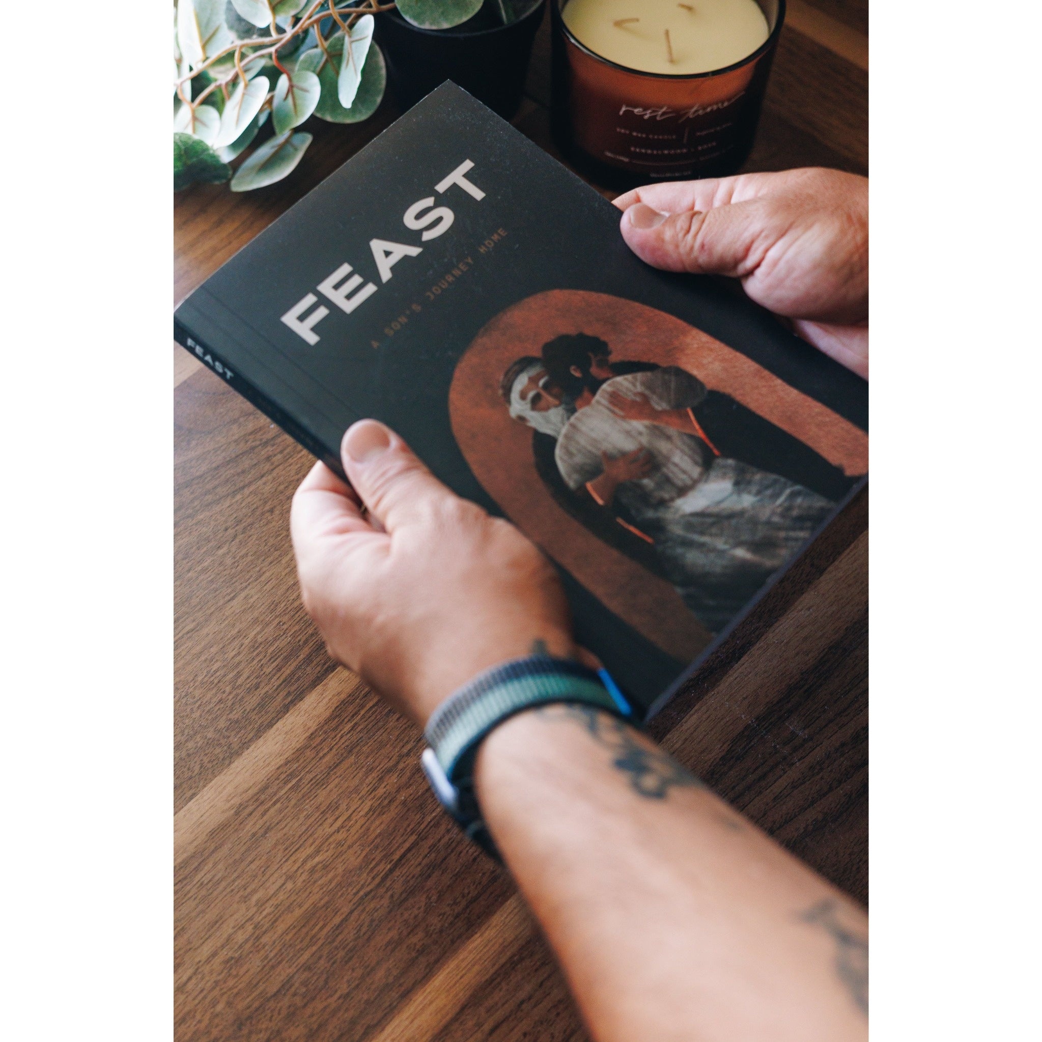 Hands hold Feast cookbook, with dark cover and illustrated image, on a wood table.