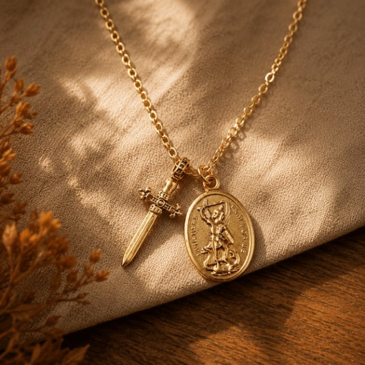 A gold-tone necklace with a round saint medallion pendant and a small sword charm, displayed on a textured beige fabric over a wooden surface with warm sunlight and dried flowers.