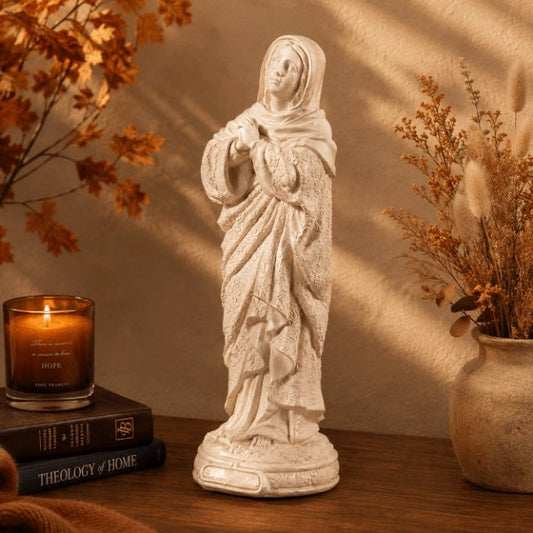 An off-white stone-look statue of the Virgin Mary stands on a wooden table in a warmly lit room. A lit amber candle on stacked books sits to the left, and a vase of dried grasses is to the right.