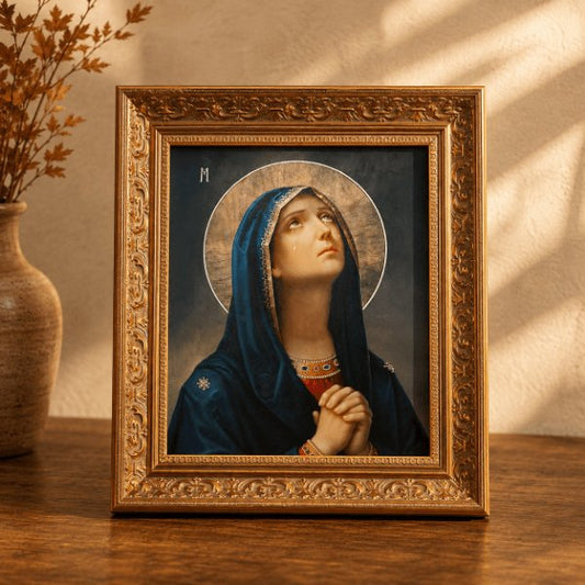 An ornate gold-framed religious art print of the Virgin Mary, styled on a wooden table next to a ceramic vase with dried branches under warm, sunlit window shadows.