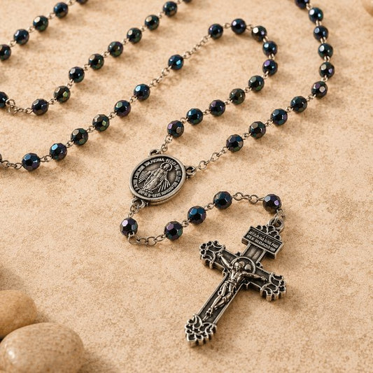 A rosary necklace with dark iridescent beads, a round medallion, and an ornate crucifix pendant, arranged on a beige textured surface with small stones.