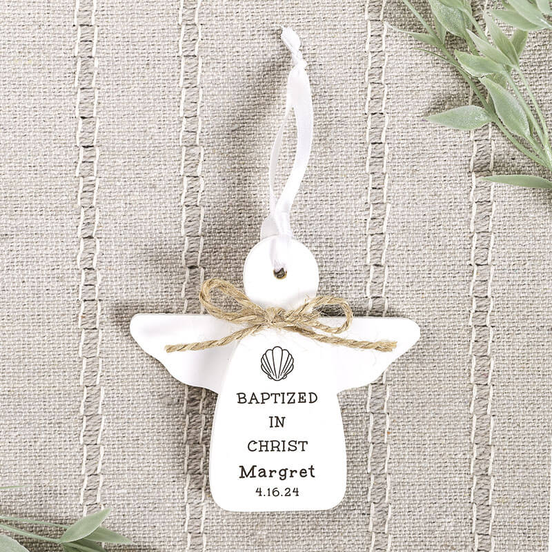 White angel-shaped ornament with 'Baptized in Christ' text on a textured fabric background.