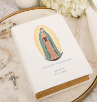 Book with Our Lady of Guadalupe illustration on a marble surface with rosary and flowers.