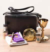Black handbag with religious items including a gold chalice and bottles on a wooden surface.