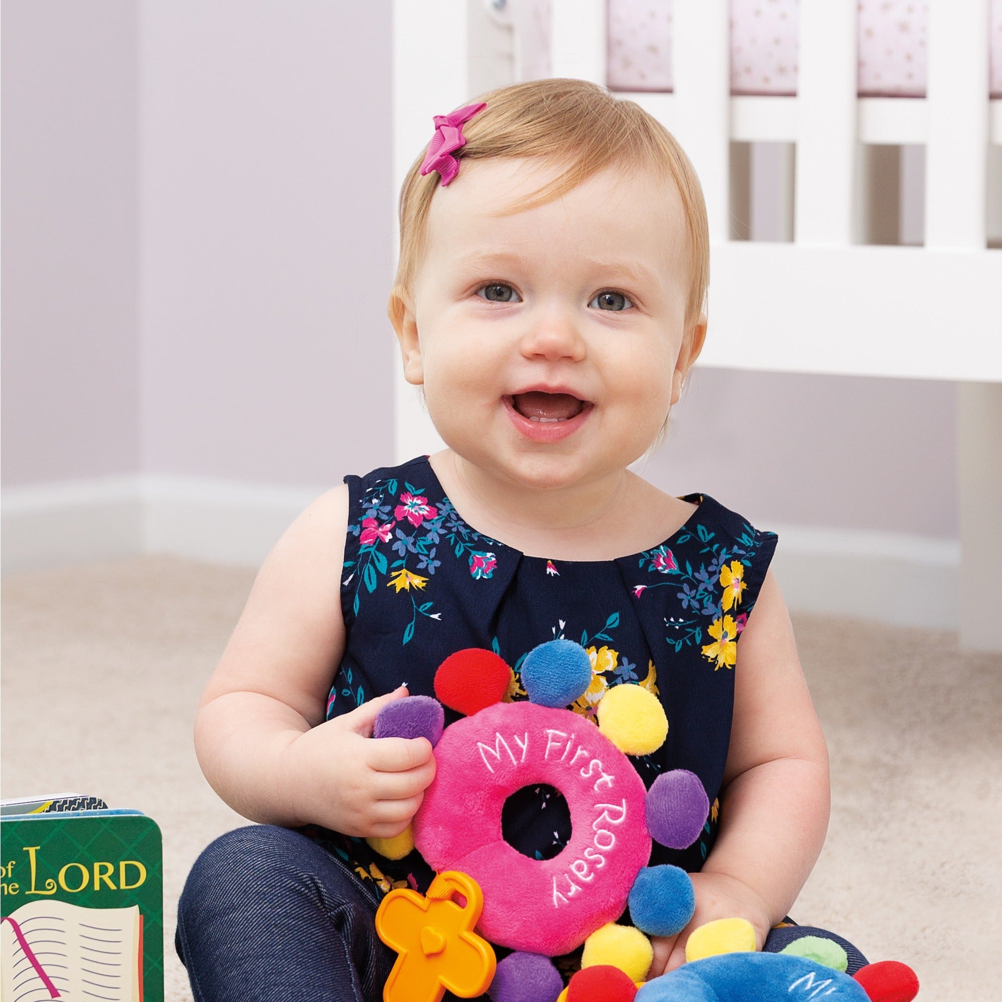 Infant holds a "My First Rosary" plush toy with colorful pom poms and a cross shape.