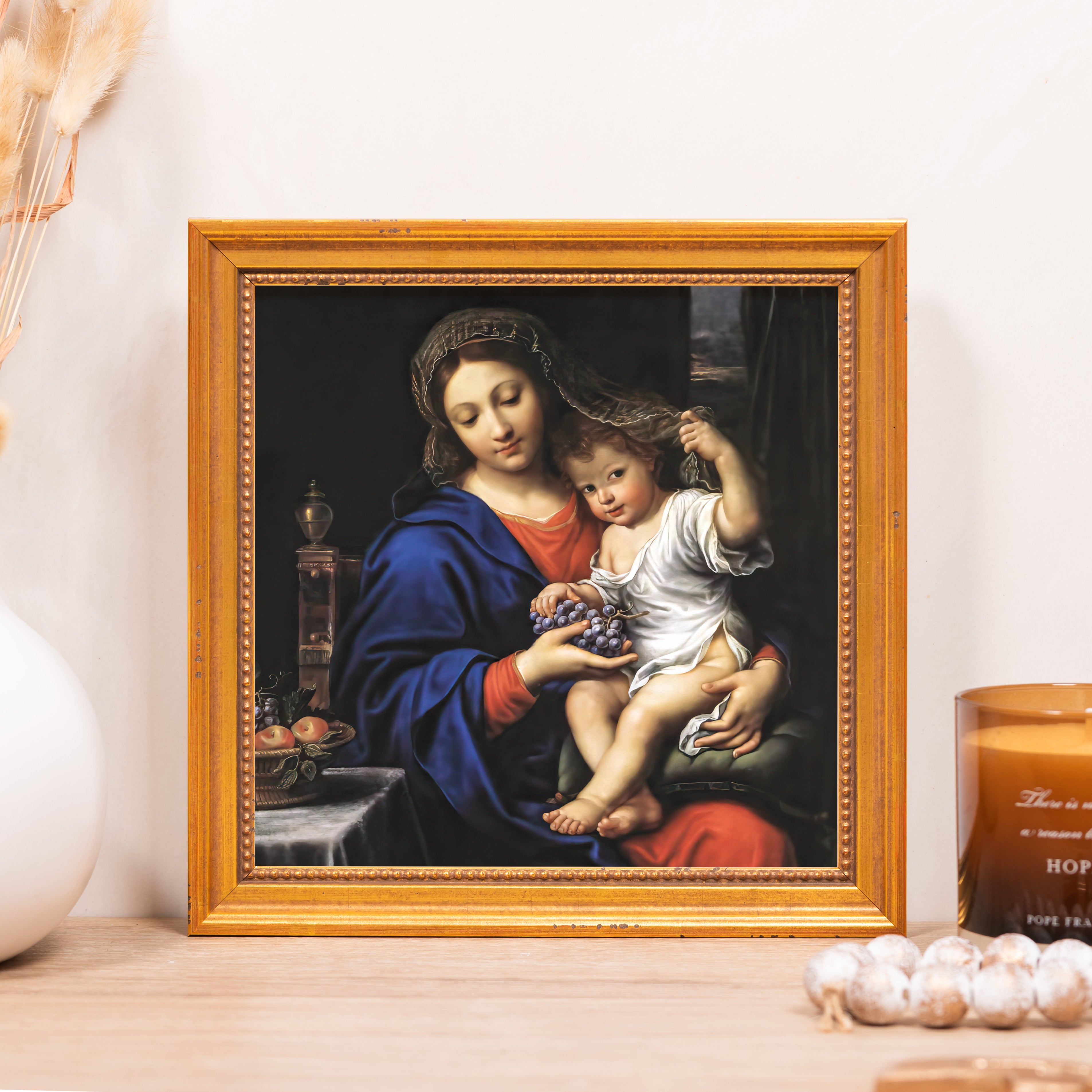 Framed religious art of Mary and child with grapes, gold frame, on table top.