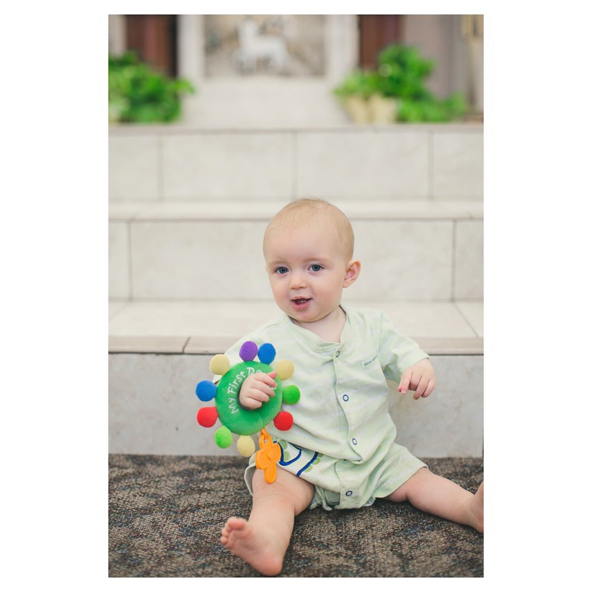 Baby holds green, fabric "My First Days" rattle toy with colorful balls attached.