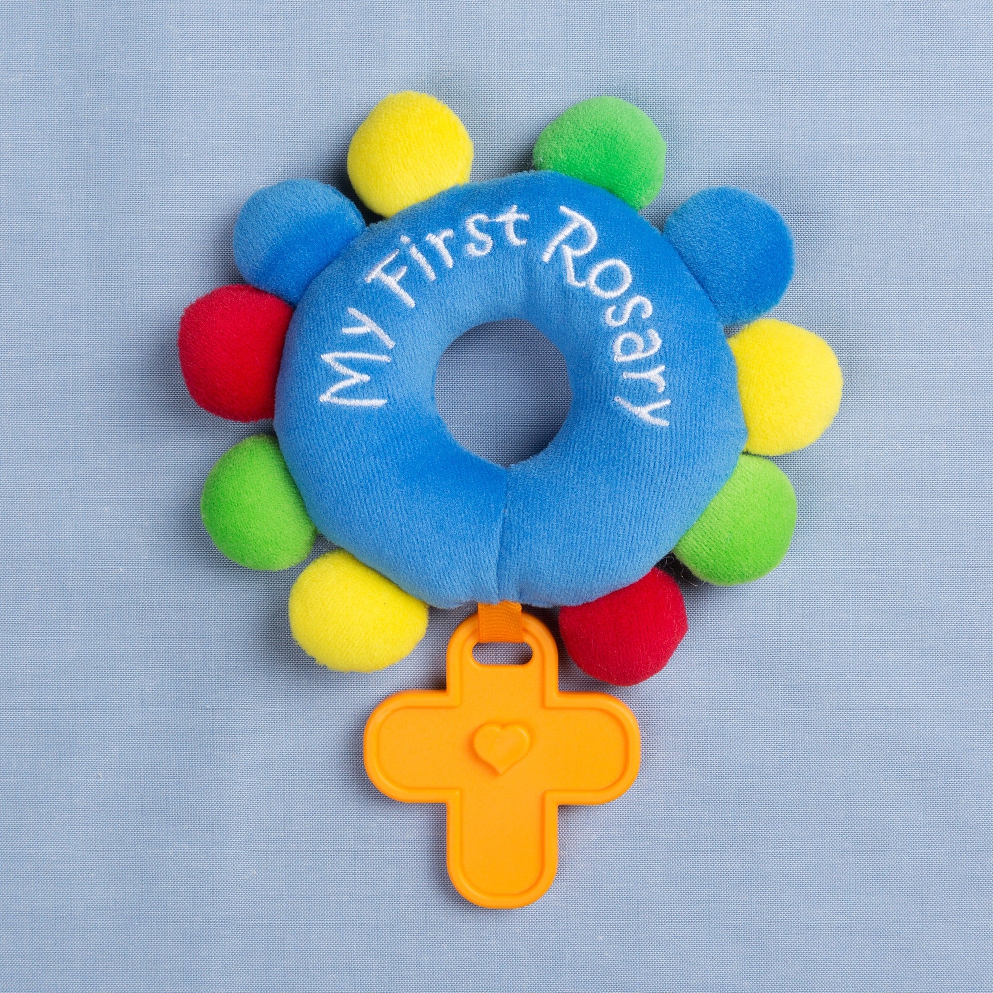Plush "My First Rosary" baby toy with blue center, colorful ball accents, and orange cross rattle.