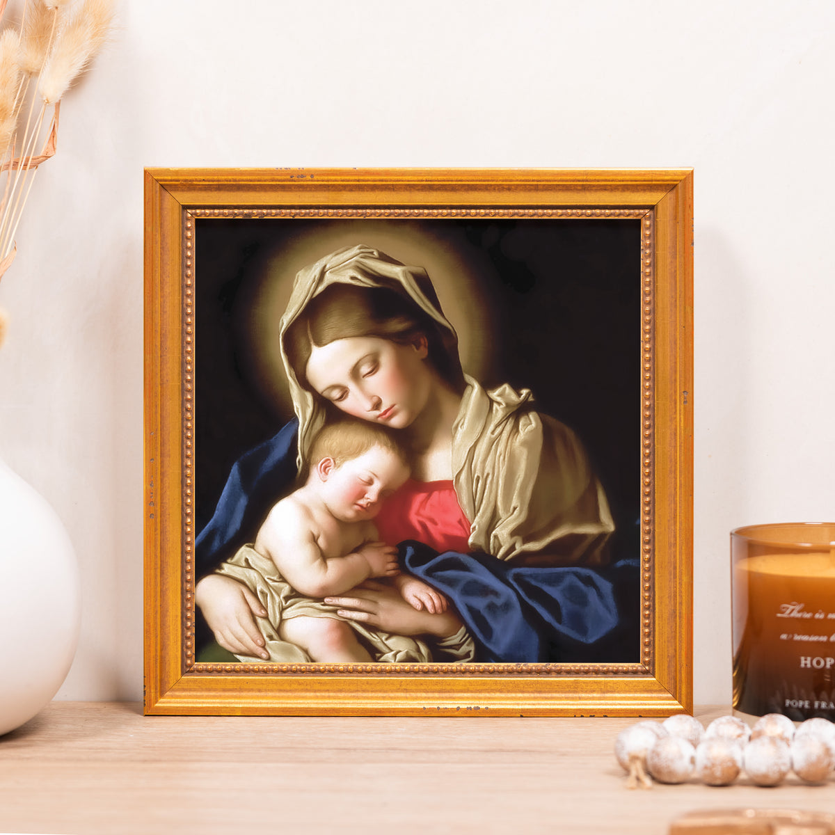 Framed painting of Mary and baby Jesus with a gold-toned frame on a light wood shelf.