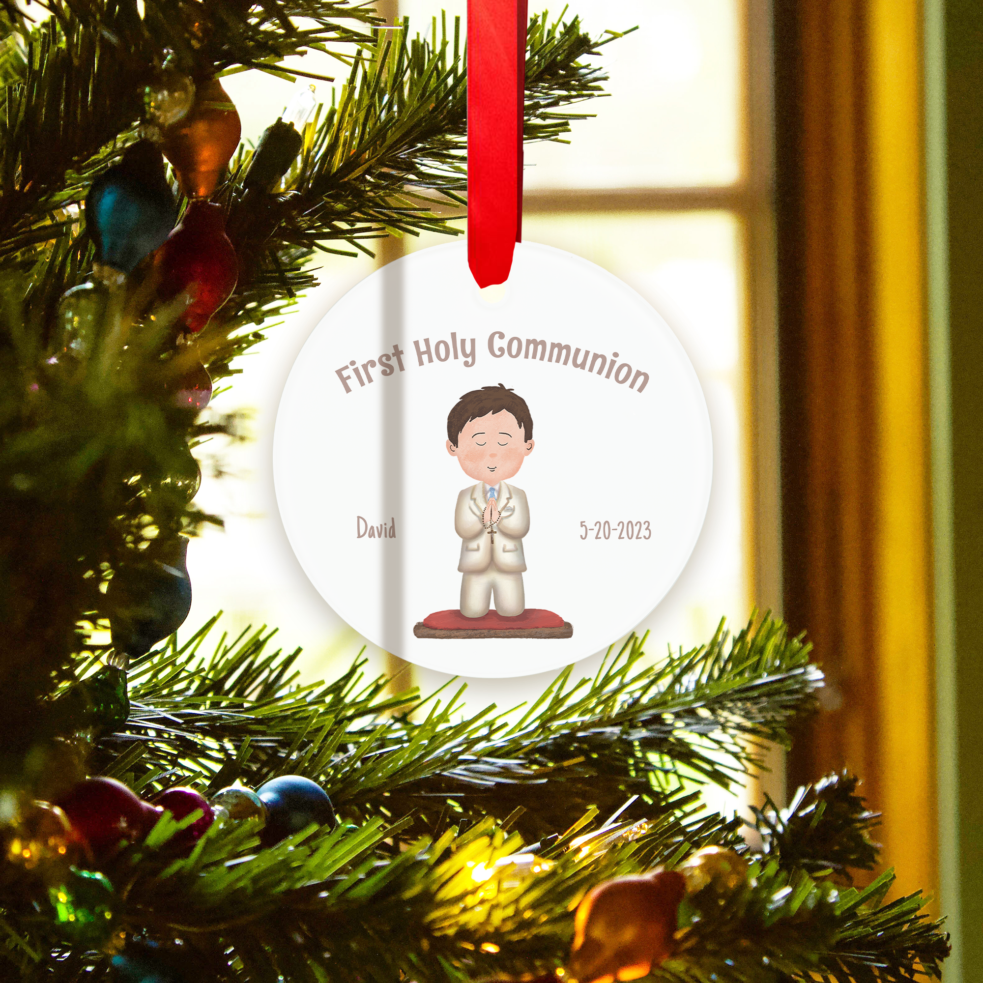 First communion ornament on tree, features a boy illustration, date, and name on a white circle with a red ribbon.