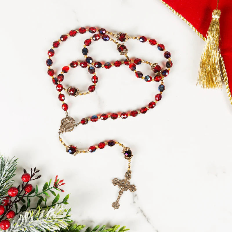 Red beaded rosary with a metal crucifix displayed on a white surface with Christmas greenery accents.