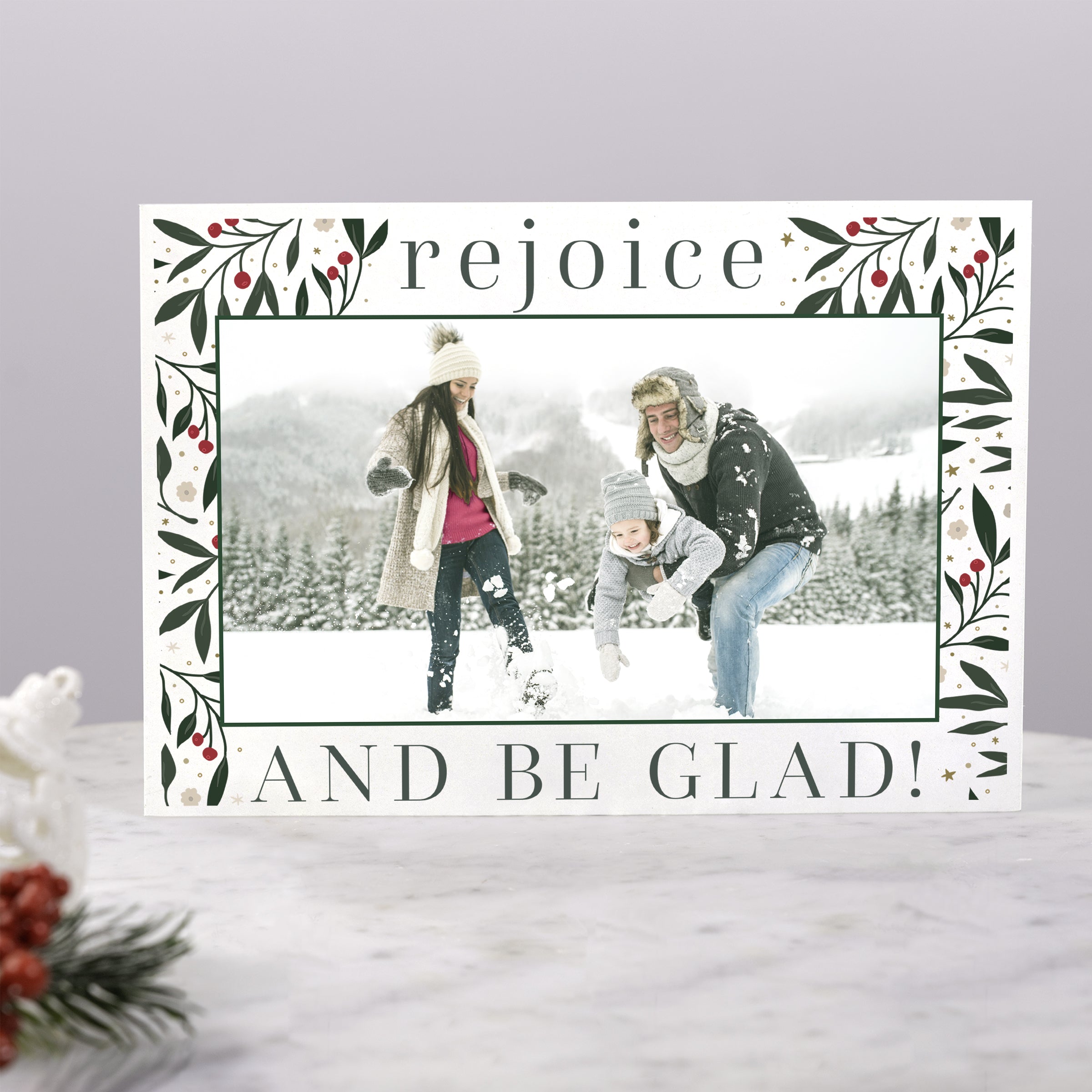 Holiday card with "Rejoice" text, family photo, and a floral design with green leaves and red berries on white paper.