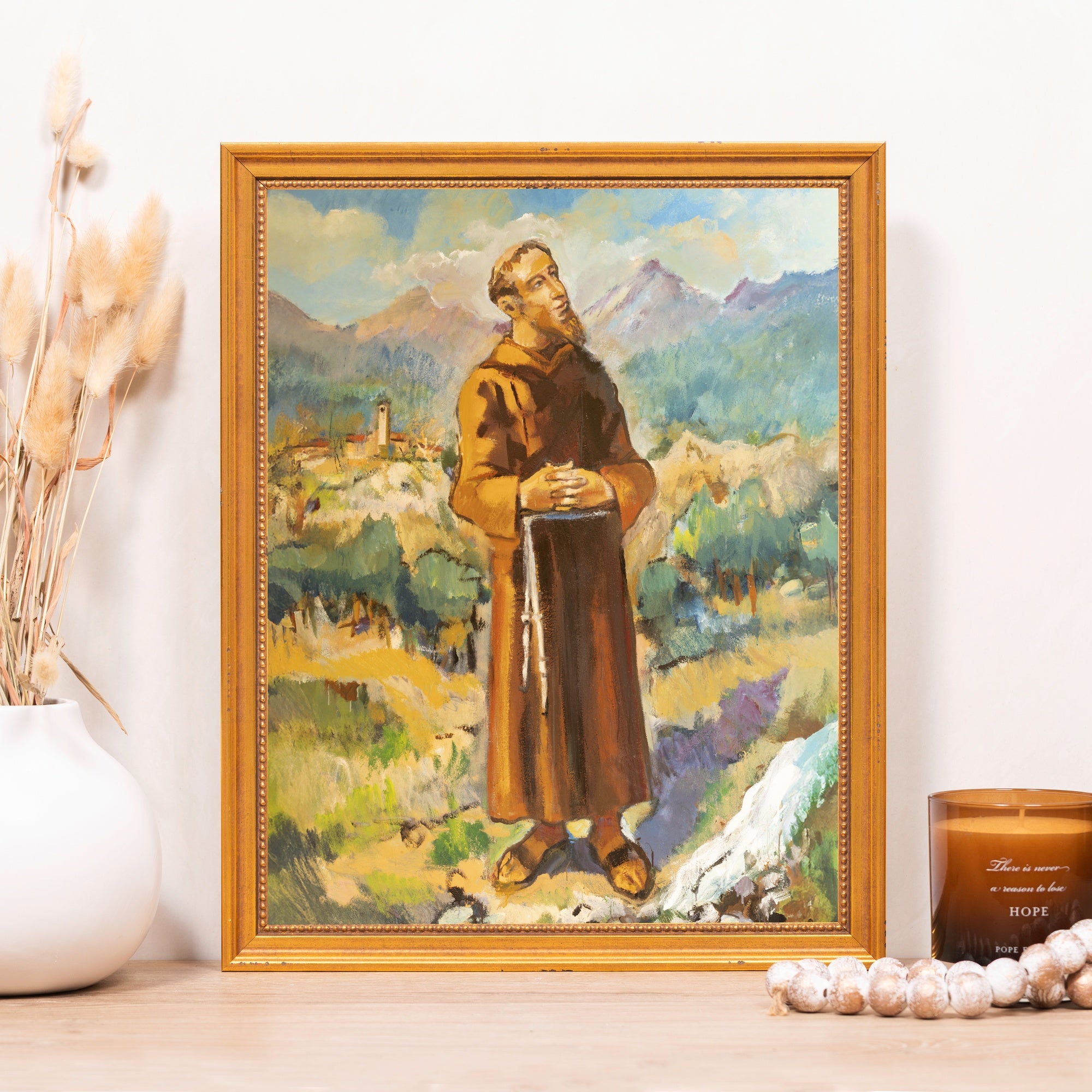 Framed oil painting of St. Francis in a brown robe and sandals, set against a background of mountains and greenery.