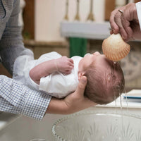 Newborn baby being held while water is poured from a shell into its hair, during a baptismal ceremony.