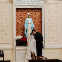 Wedding couple kneeling in front of a statue of Mary in a church.