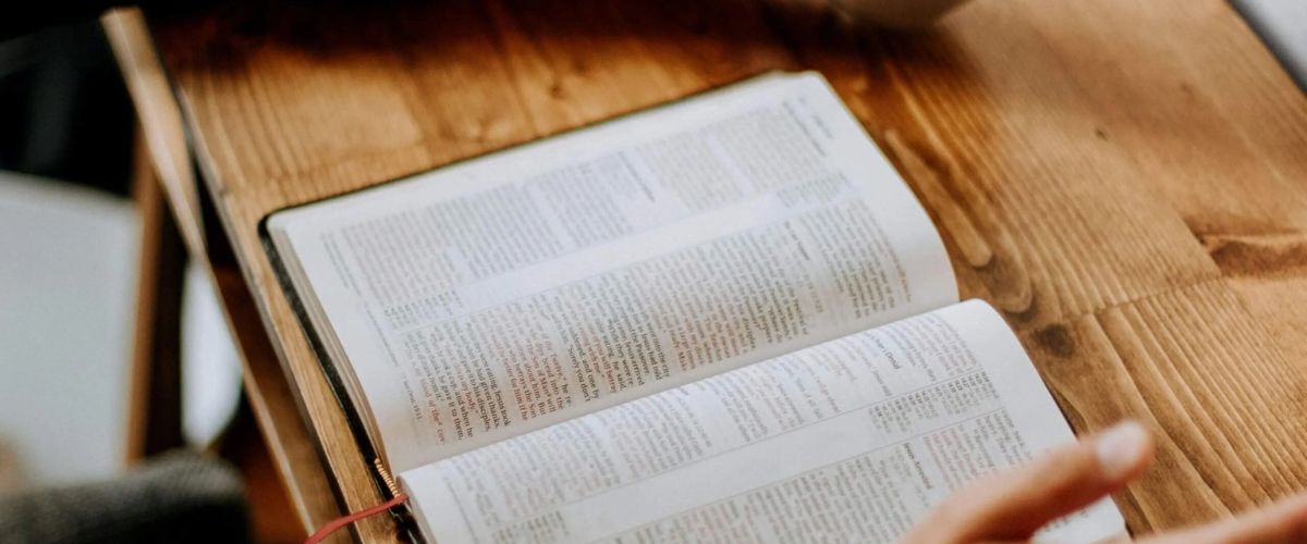 Hands holding a coffee cup over an open Bible on a wooden table, suggesting quiet reading or devotional time.
