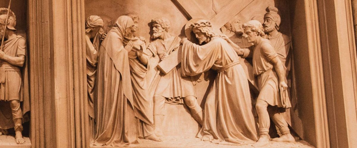 Stone relief showing Jesus carrying the cross while meeting his mother, surrounded by figures, from the Stations of the Cross.