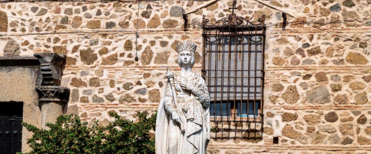Statue of Isabella I of Castile standing before a rustic stone wall with a barred window
