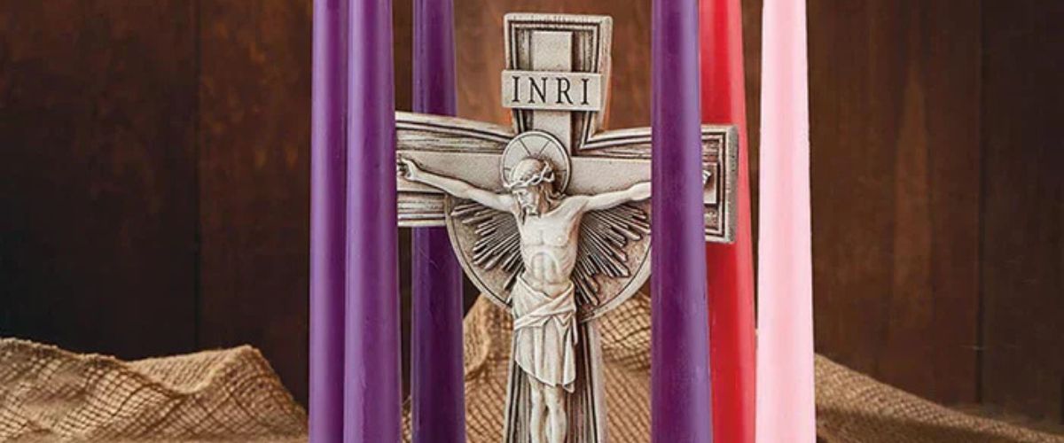 **Alt text:** Crucifix with six lit candles in purple, red, and pink arranged around it on a rustic background.