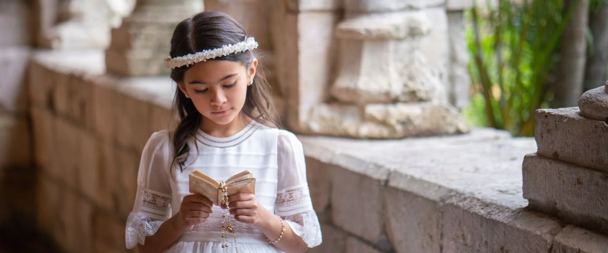 Young girl in a white dress and floral headband holding a small prayer book and rosary in a church setting
