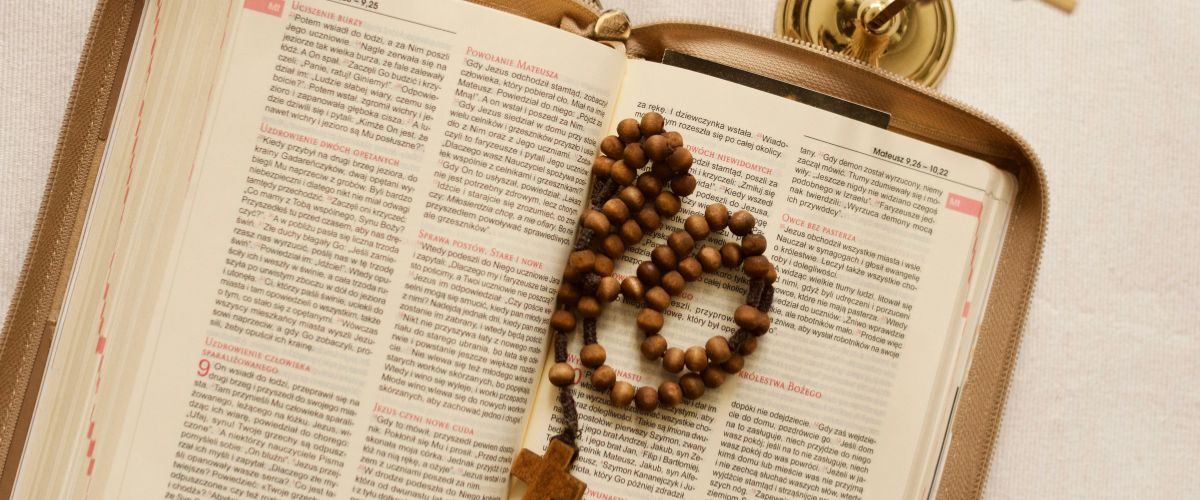 Wooden rosary with crucifix resting on an open Bible beside a lit candle.