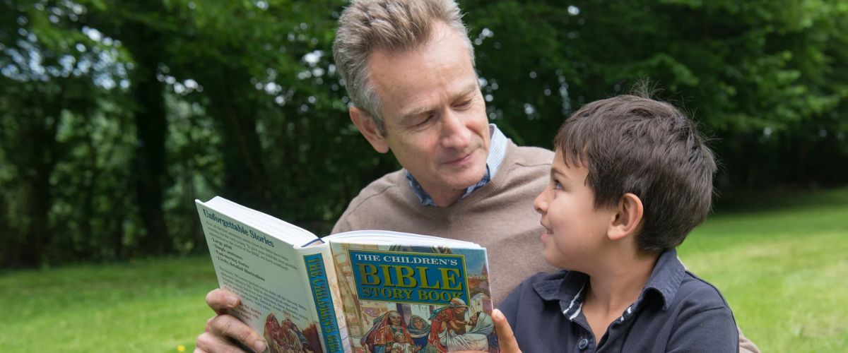 Man reading a children’s Bible storybook with a young boy outdoors.