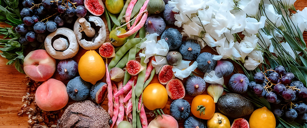 Fresh assortment of fruits, vegetables, mushrooms, grains, and white flowers arranged on a wooden table
