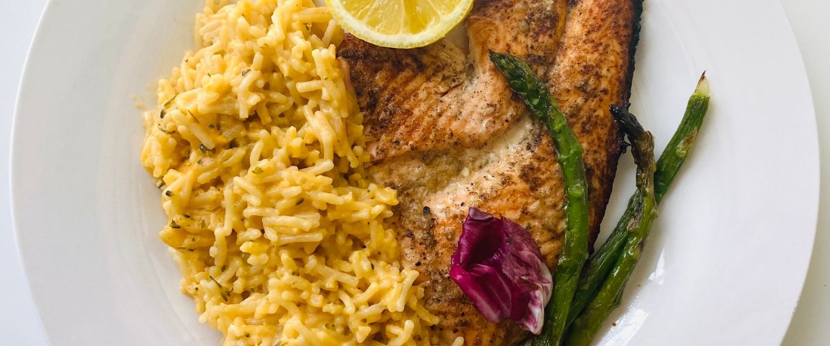 Grilled salmon fillet with lemon slice, creamy rice, and roasted asparagus served on a white plate.