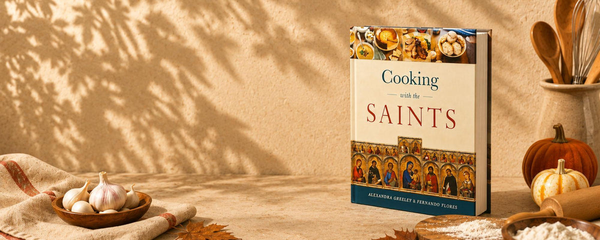 The cookbook "Cooking with the Saints" by Alexandra Greeley & Fernando Flores displayed on a warm, autumn-themed kitchen counter with pumpkins, garlic, and baking utensils.