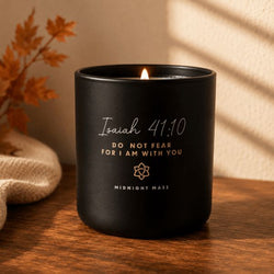 A black candle jar with a lit flame sits on a wooden surface. The front is printed with the text 'Isaiah 44:10 DO NOT FEAR FOR I AM WITH YOU' and 'MIDNIGHT MASS' in white and gold lettering.