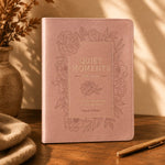 A pink faux-leather journal with embossed floral borders and the title 'QUIET MOMENTS FOR MOMS' on the cover, styled on a wooden table with warm sunlight, a vase, folded fabric, and a pen.