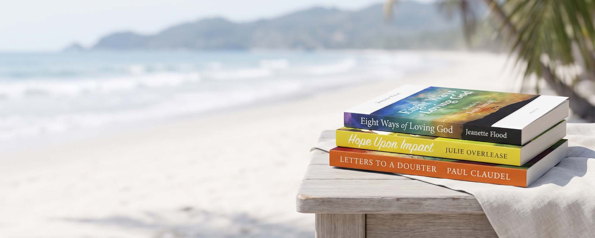 Three paperback books stacked on a white wooden table outdoors on a beach, with a blurred ocean and shoreline in the background. The book spines and covers show titles including 'Eight Ways of Loving God', 'Hope Through Impact', and 'Letters to a Bouter' by Jennifer Hood and Paul Claudel.