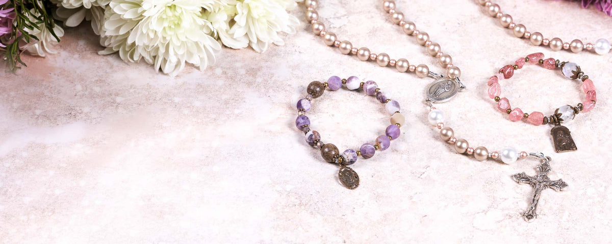 Rosary bracelets and rosaries in elegant pinks and purples displayed with a floral backdrop.