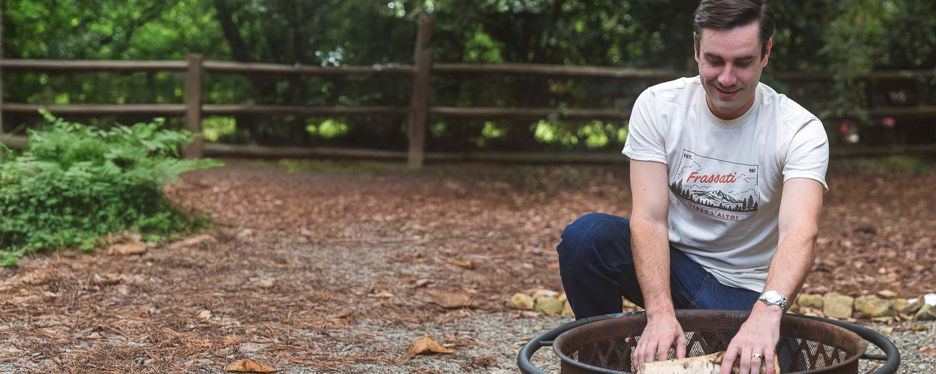 Man kneeling outdoors, smiling as he arranges items in a metal fire pit.