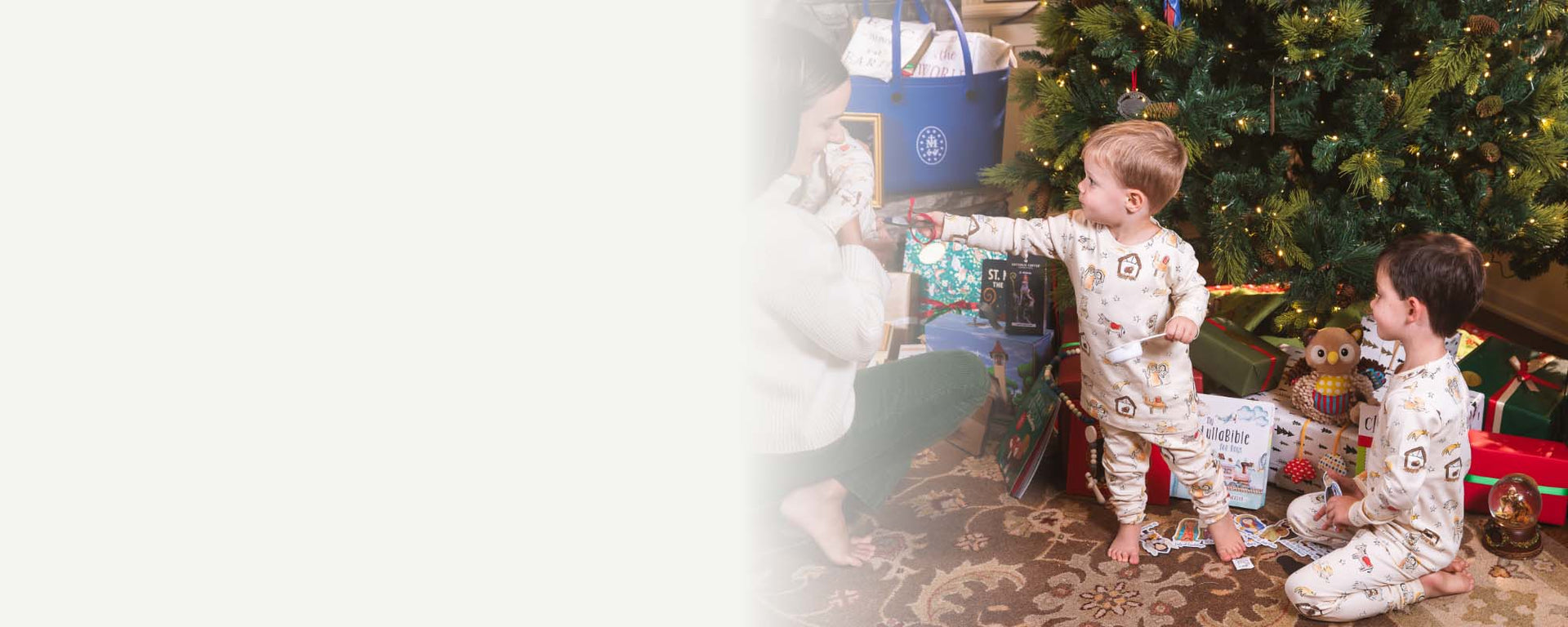 Two young children in matching pajamas open gifts by a decorated Christmas tree while an adult kneels nearby, smiling
