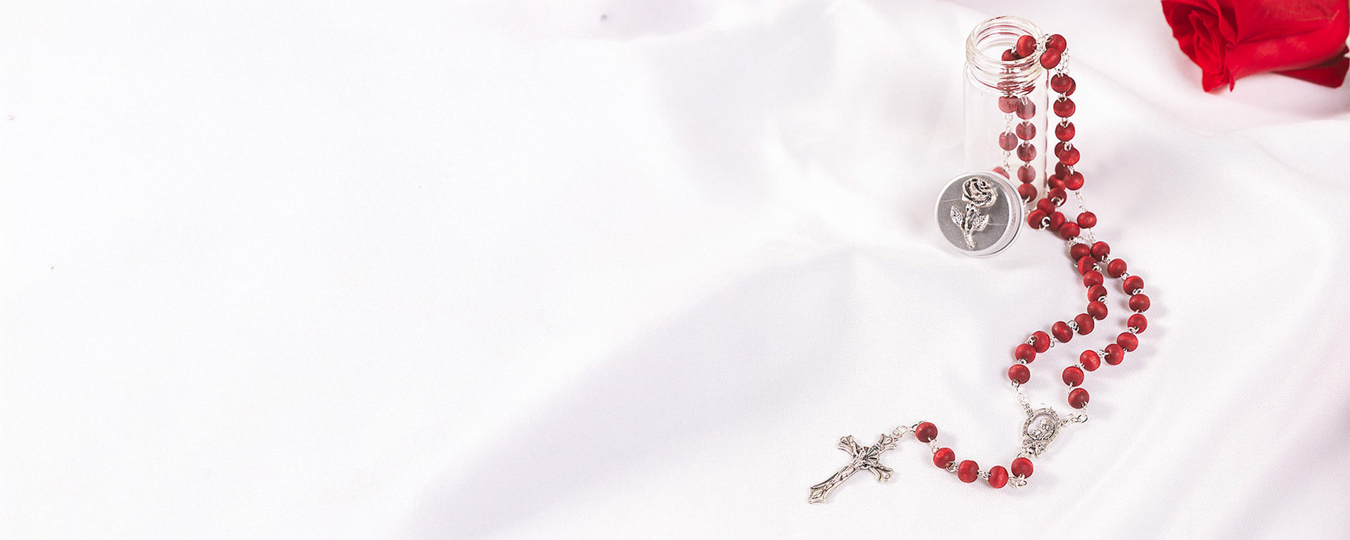 Red rosary beads with a silver crucifix and rose medallion in a small glass bottle on a white background, with a red rose in the corner.