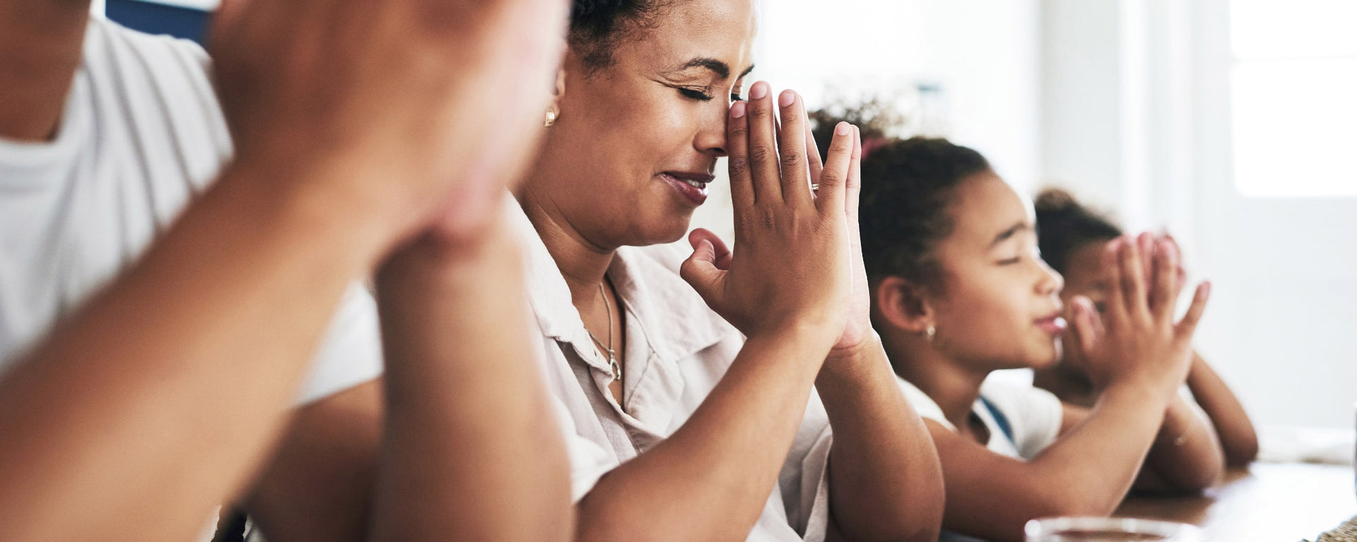 A family of four praying together in a brightly lit room. The mother is in the foreground with her hands clasped together, smiling, followed by her children behind her.